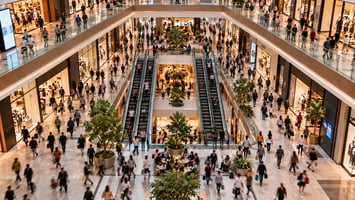 **ALT text:** “Busy multi-level shopping centre with visitors moving between stores, escalators and shared spaces.”