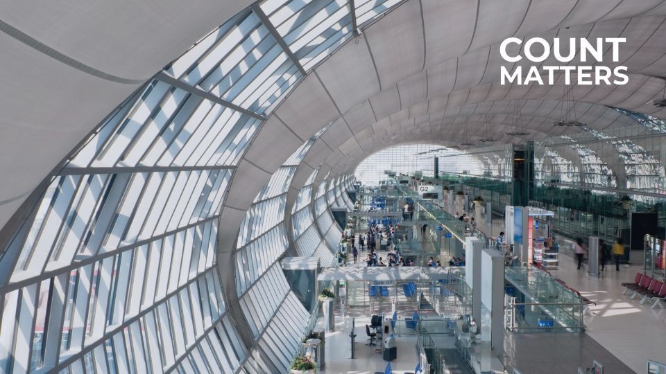 Interior of Bangkok Suvarnabhumi Airport with curved glass ceiling, showing passengers walking through the terminal and CountMatters logo overlayed in the corner.