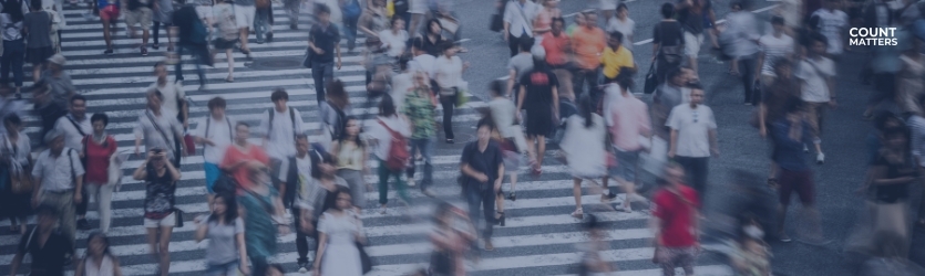 Sløret menneskemængde, der krydser et stort bykryds og repræsenterer en kraftig fodgængerstrøm. Ideel til at illustrere fodgængeranalyser, overvågning af fodgængertrafik og smart city-mobilitetsplanlægning med CountMatters, logo i hjørnet.