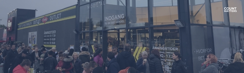 Travl outlet-butik med stor menneskemængde udenfor og fed OUTLET-skiltning. Ideel til at illustrere fodfaldsanalyser i detailhandlen, overvågning af besøgende i spidsbelastningsperioder og konverteringsoptimering med CountMatters, logo i hjørnet.