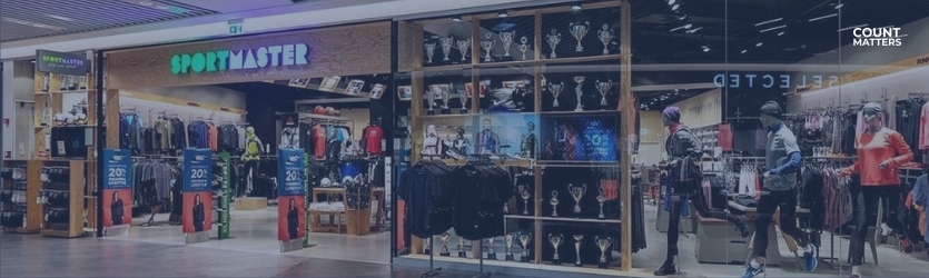 Indgang til Sportmaster-butik med mannequiner, udstilling af sportstøj og reklameskilte. Ideel til at illustrere detailhandelsanalyser, besøgstælling og optimering af konverteringsfrekvens med CountMatters, logo i hjørnet.