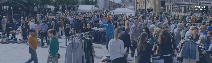 Travlt udendørs marked på et torv i byen med masser af mennesker, der handler i tøjboder. Ideel til at illustrere besøgsanalyser, overvågning af fodfald i detailhandlen og optimering af det offentlige rum med CountMatters, logo i hjørnet.